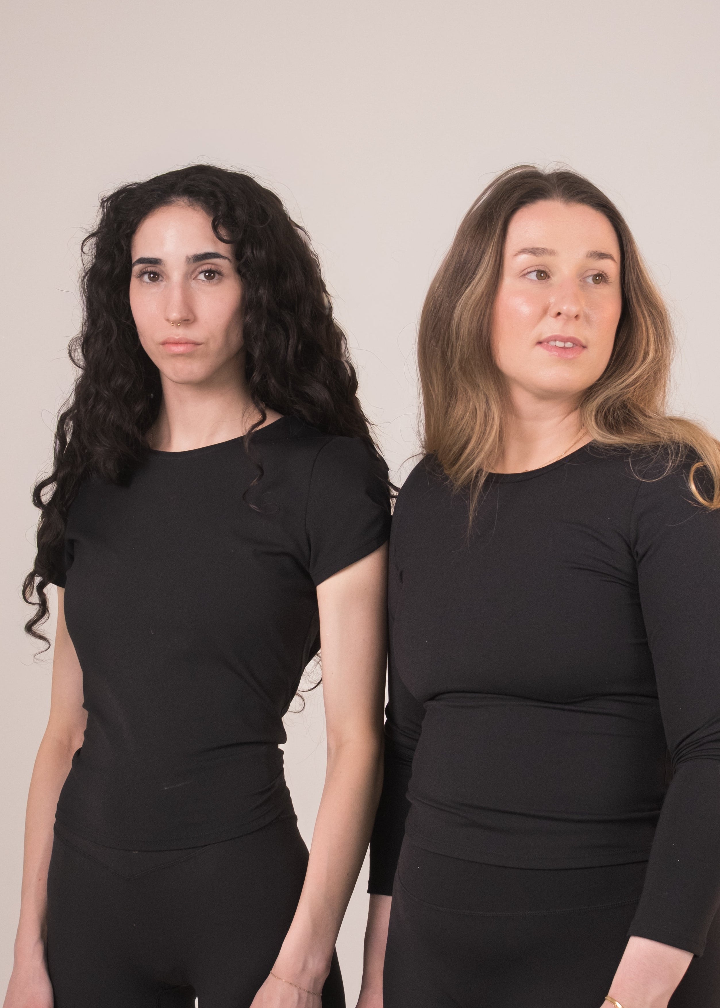 Two women wearing black outfits against a plain background