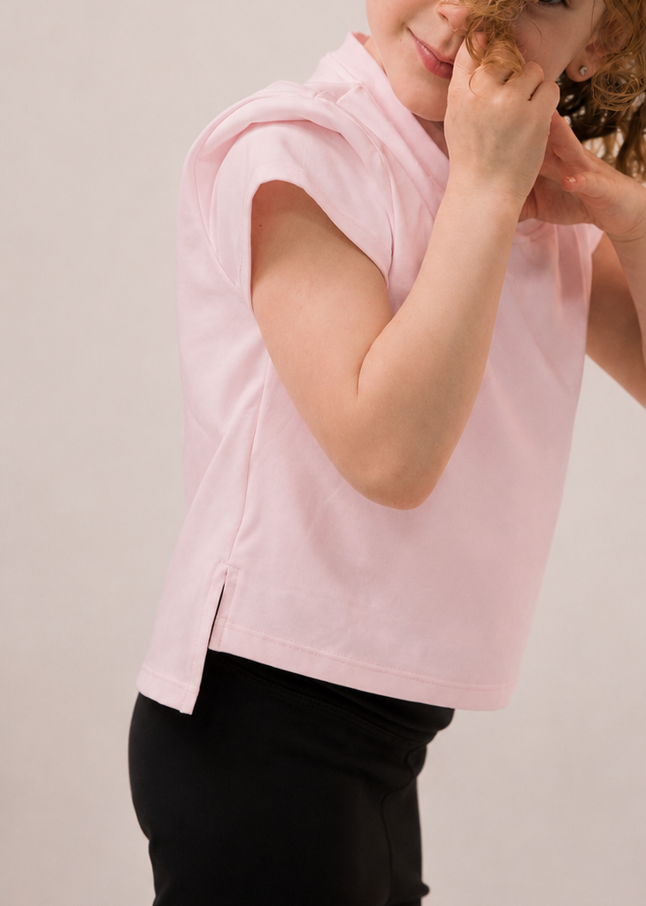 Young girl in a pink sleeveless top and black pants posing with one hand on her face against a plain background